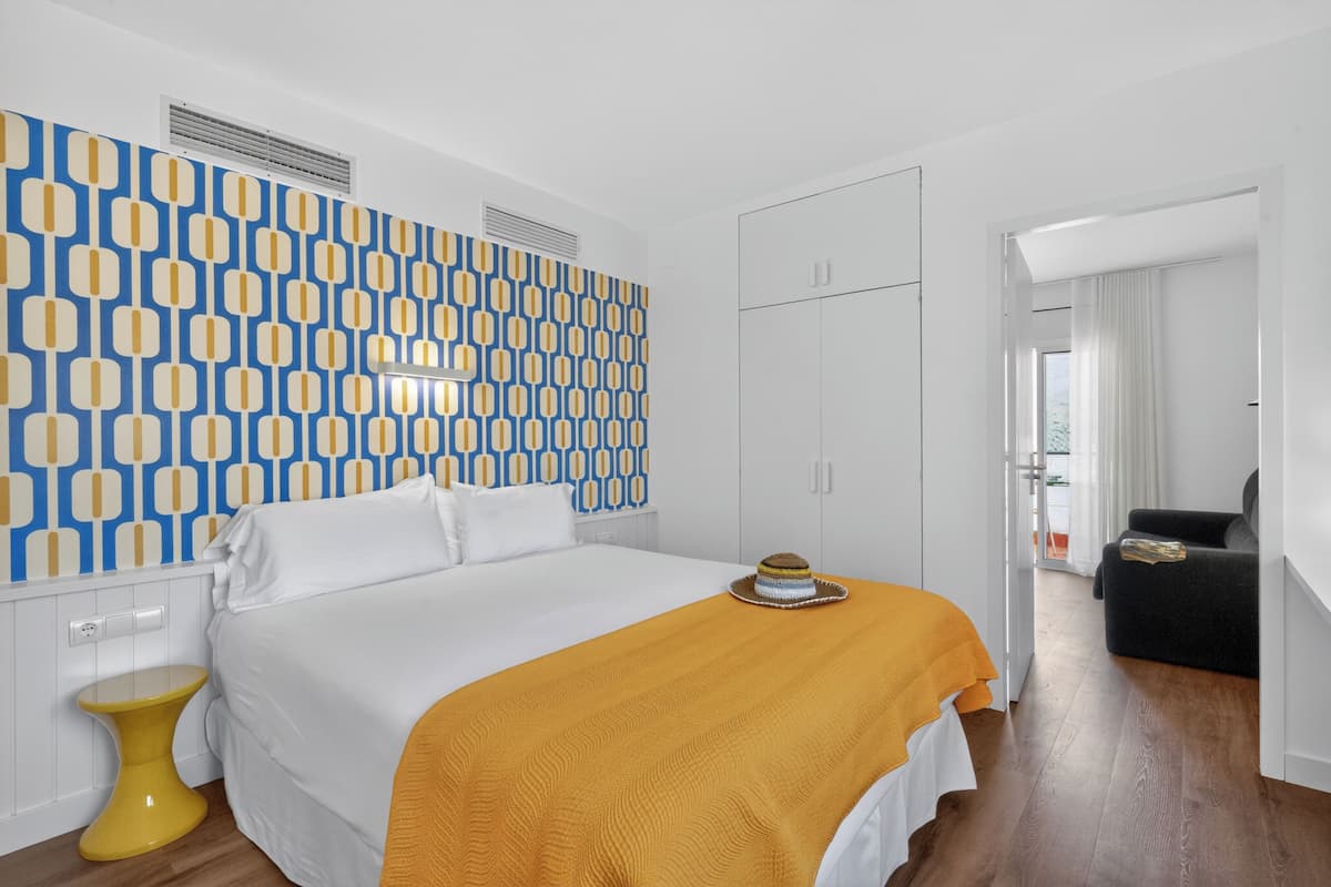 Hotellihuone, jossa on kuvioitu seinä, keltainen peitto sängyllä, pieni keltainen jakkara ja musta sohva.