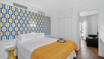 Una habitación de hotel con un papel tapiz estampado, una cama con una manta amarilla, un taburete amarillo pequeño y un sofá negro.
