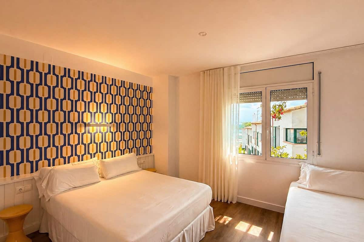 Hotellihuone, jossa on kaksi sänkyä, kuviollinen seinä, verhoilla varustettu ikkuna ja näkymä ulkona oleviin rakennuksiin.