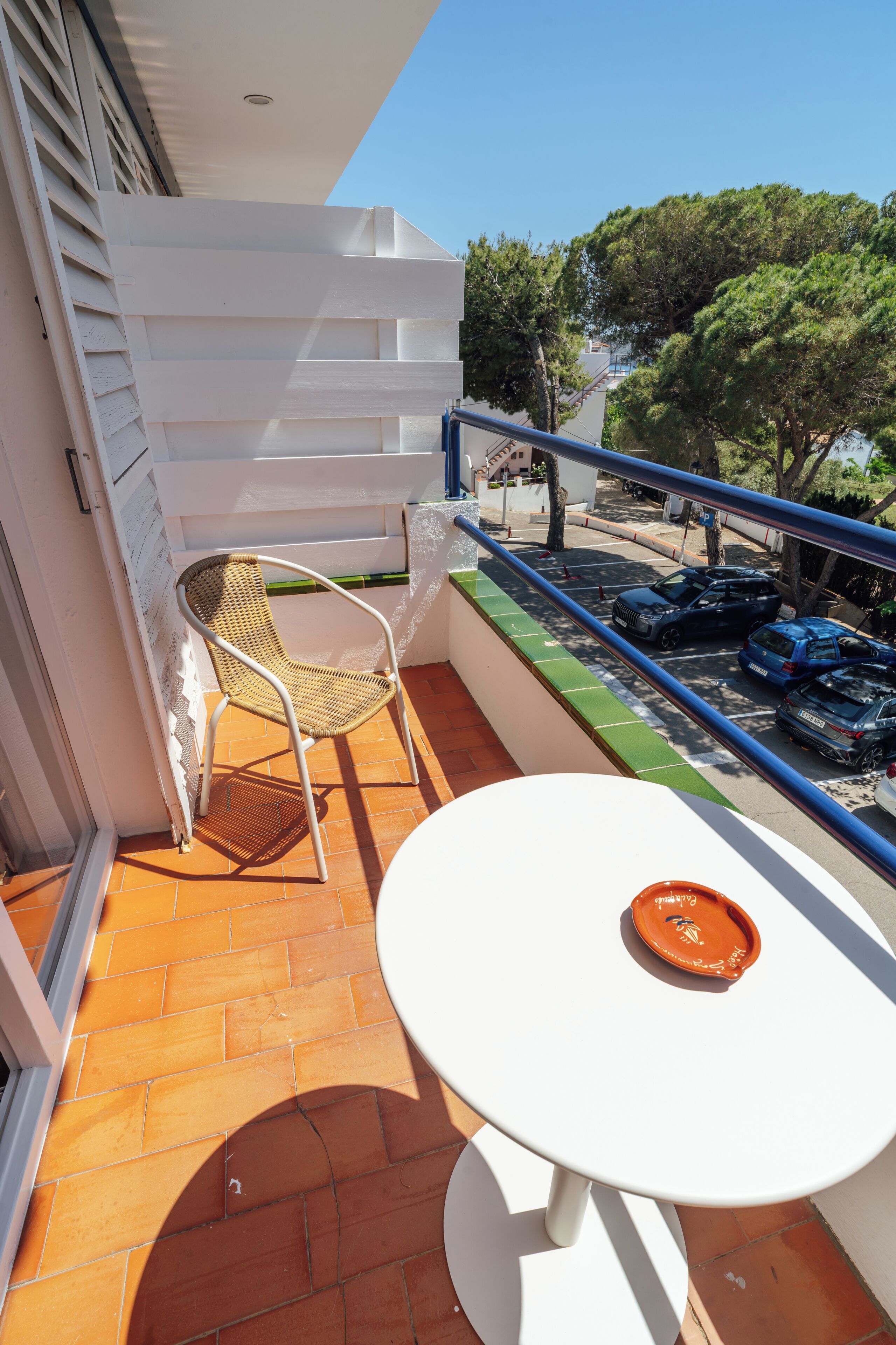 Un balcón con una mesa y silla blancos, suelo de baldosas y vista a coches aparcados y árboles.