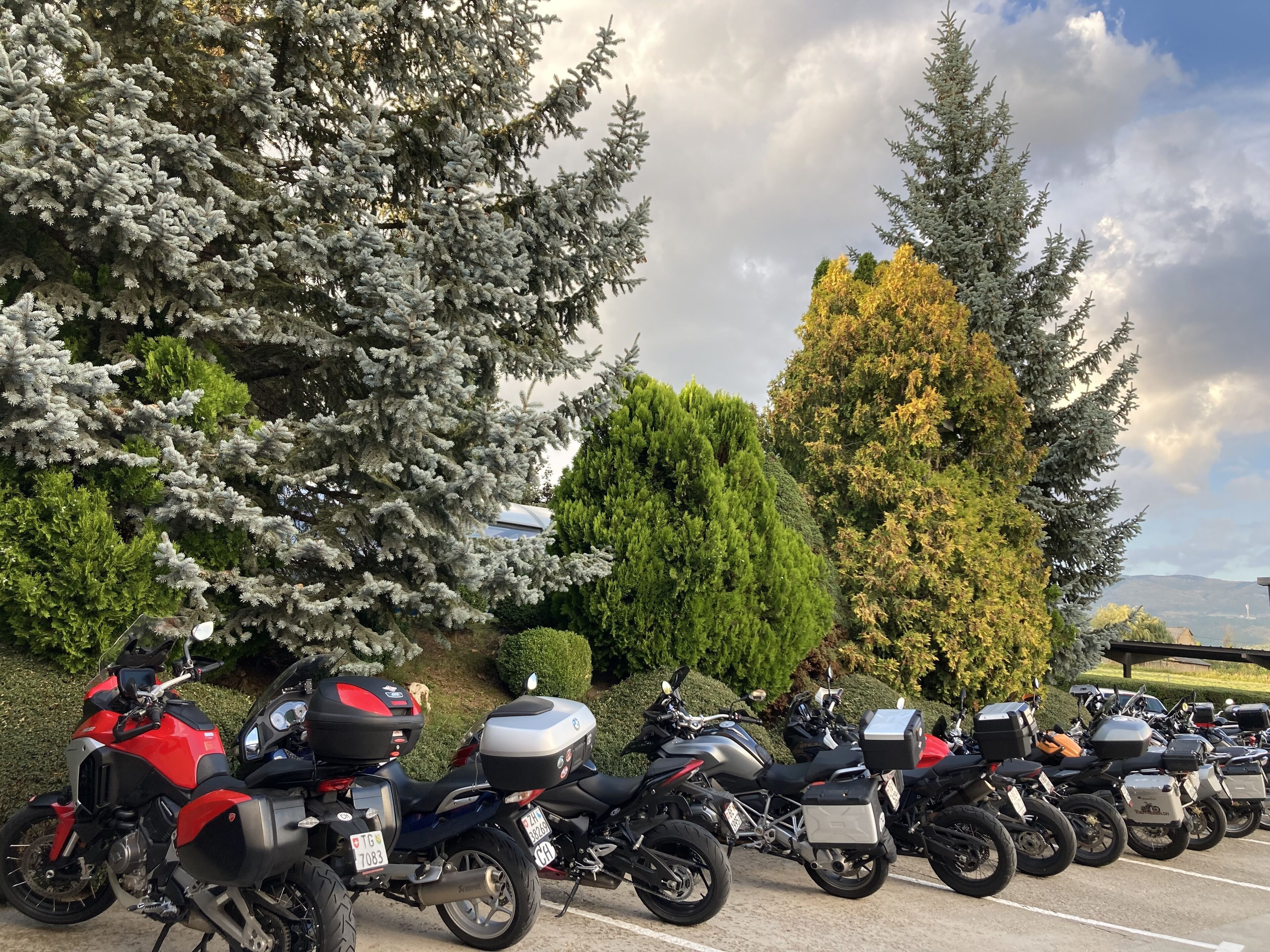 Eine Reihe von Motorrädern steht auf einem Parkplatz, im Hintergrund Bäume und ein bewölkter Himmel.