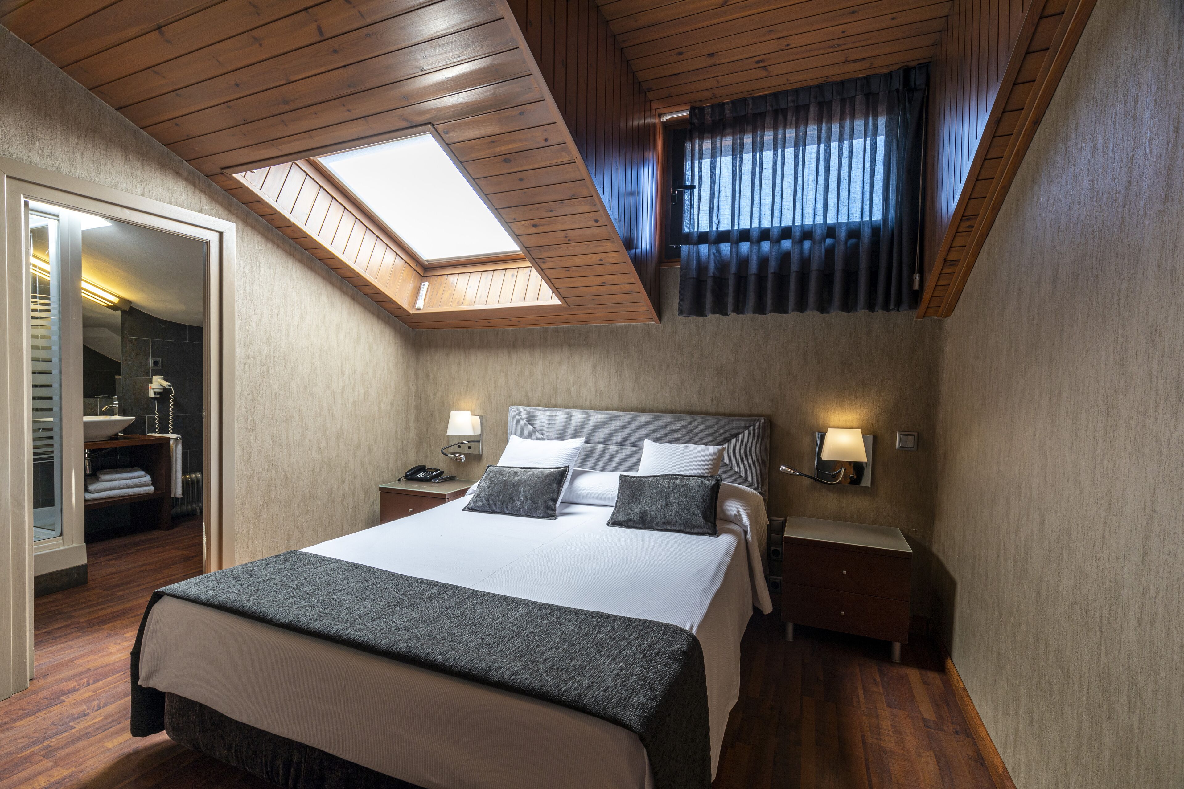 Un dormitorio con tragaluces, una cama con almohadas, una mesita de noche, una lámpara y un baño visible a través de una puerta abierta.