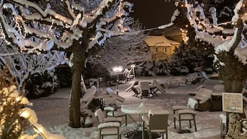 A snow-covered outdoor seating area with tables and chairs, surrounded by trees and a building in the background.