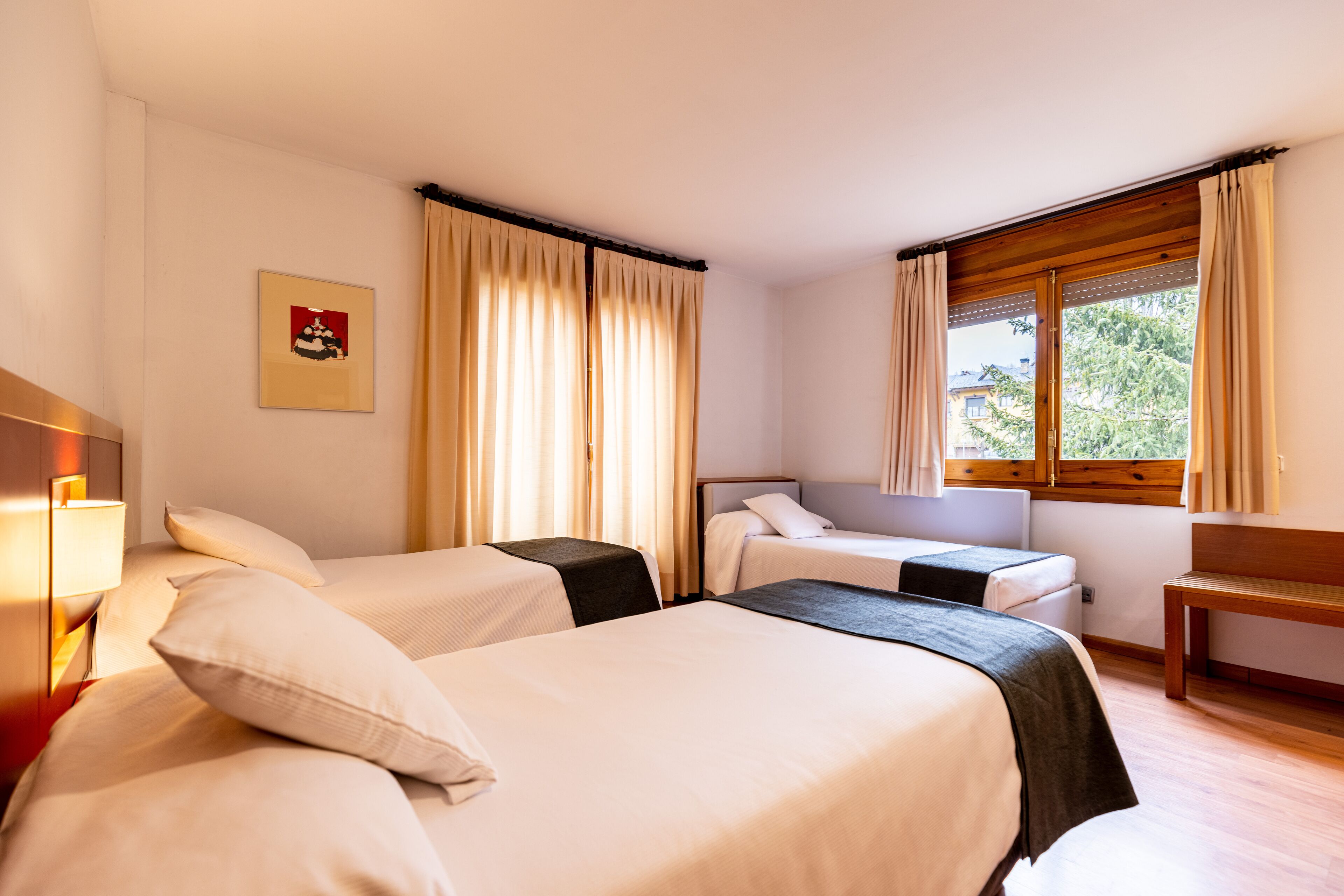 Quarto de hotel com duas camas, um banco de madeira, uma escrivaninha e uma janela com cortinas.