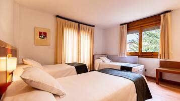 Quarto de hotel com duas camas, um banco de madeira, uma escrivaninha e uma janela com cortinas.