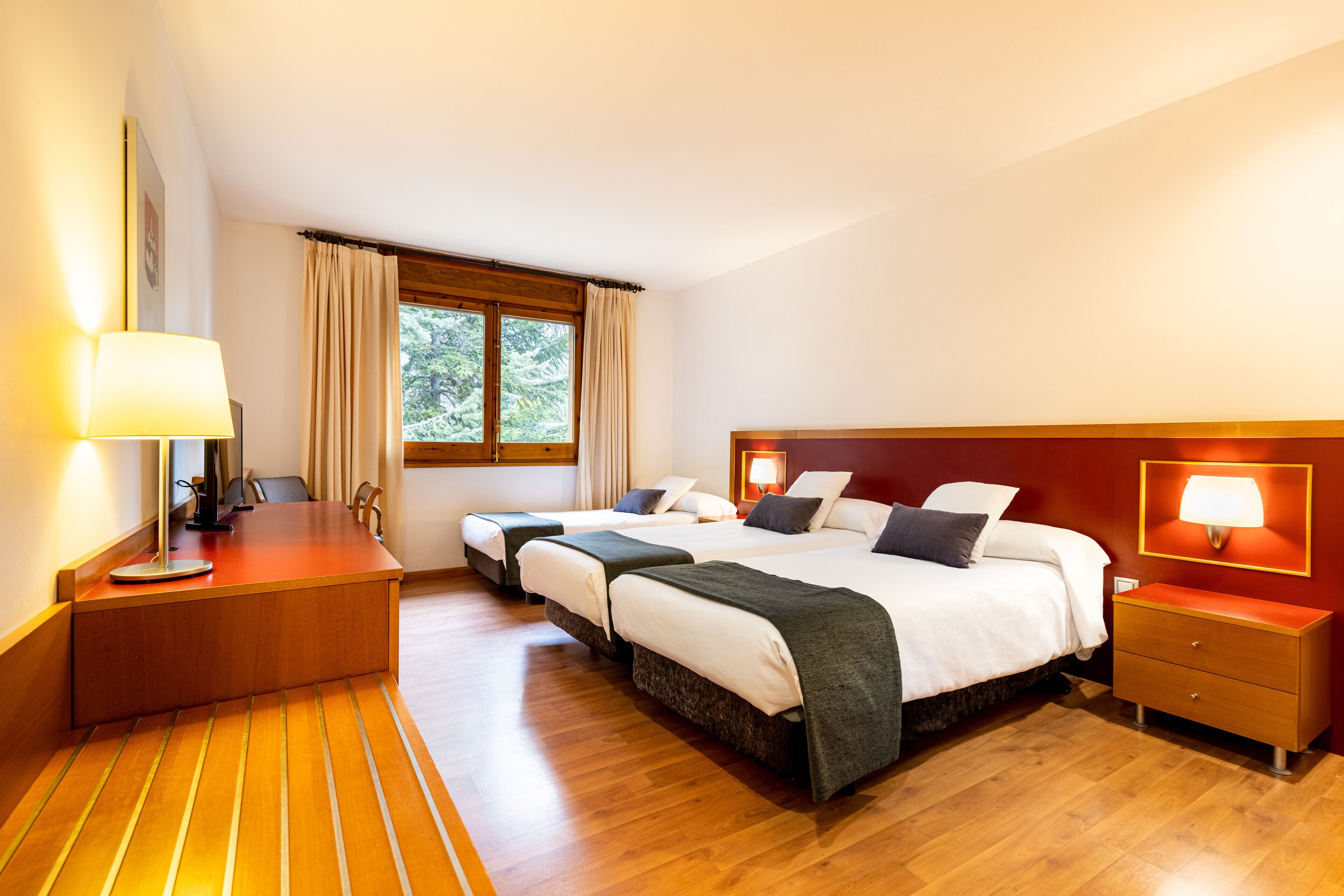 A hotel room with a wooden floor, a desk with a lamp, two beds with headboards, a nightstand, and a window with curtains.
