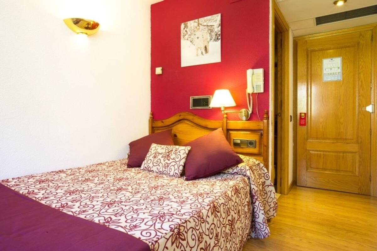 Un dormitorio con cama, una mesita de noche con lámpara, un teléfono en la pared y una puerta.