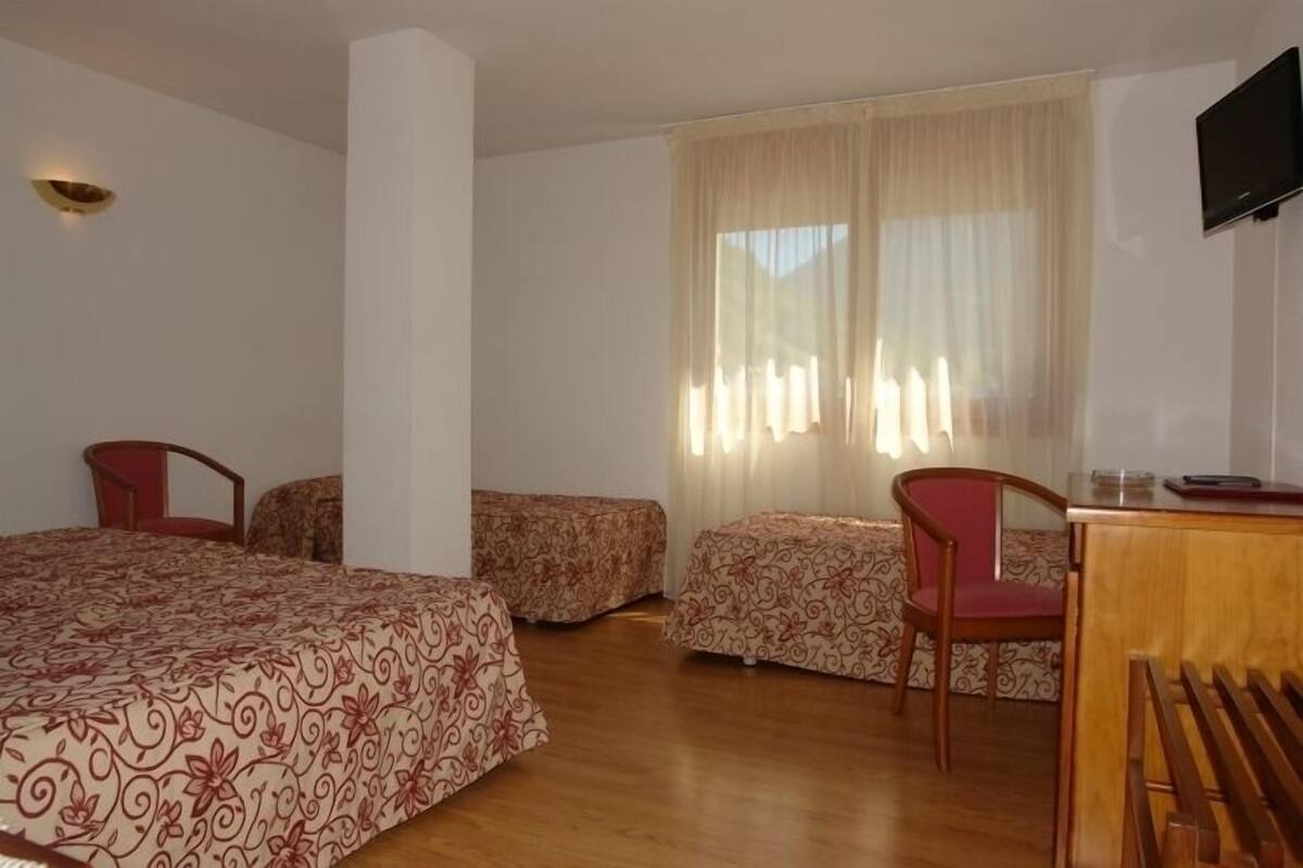 Habitación de hotel con una cama grande, dos camas más pequeñas, un escritorio con una silla, un televisor montado en la pared y una ventana con cortinas.