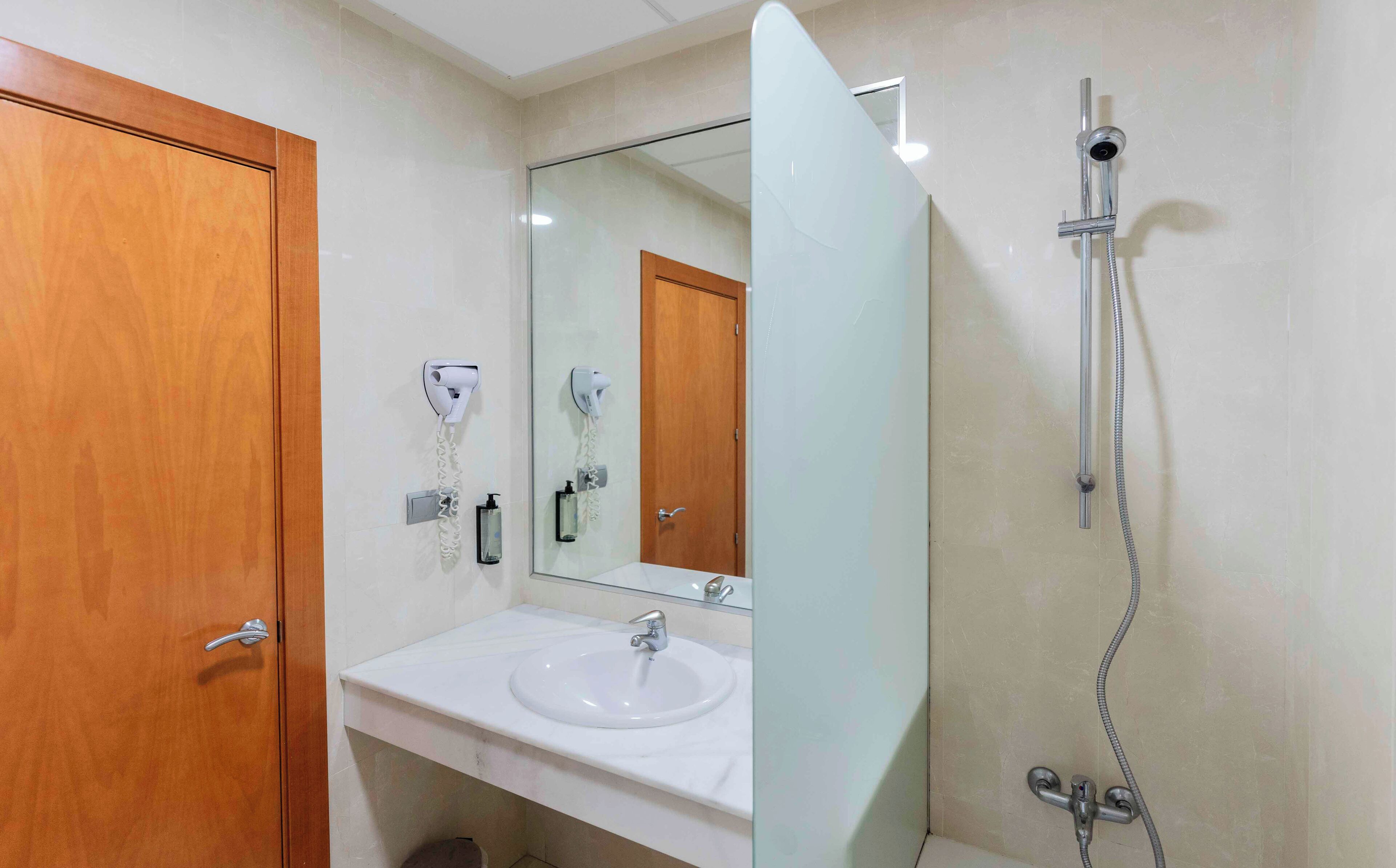 Un baño con puerta de madera, un lavamanos blanco, un espejo y una ducha con cabezal de ducha manual.