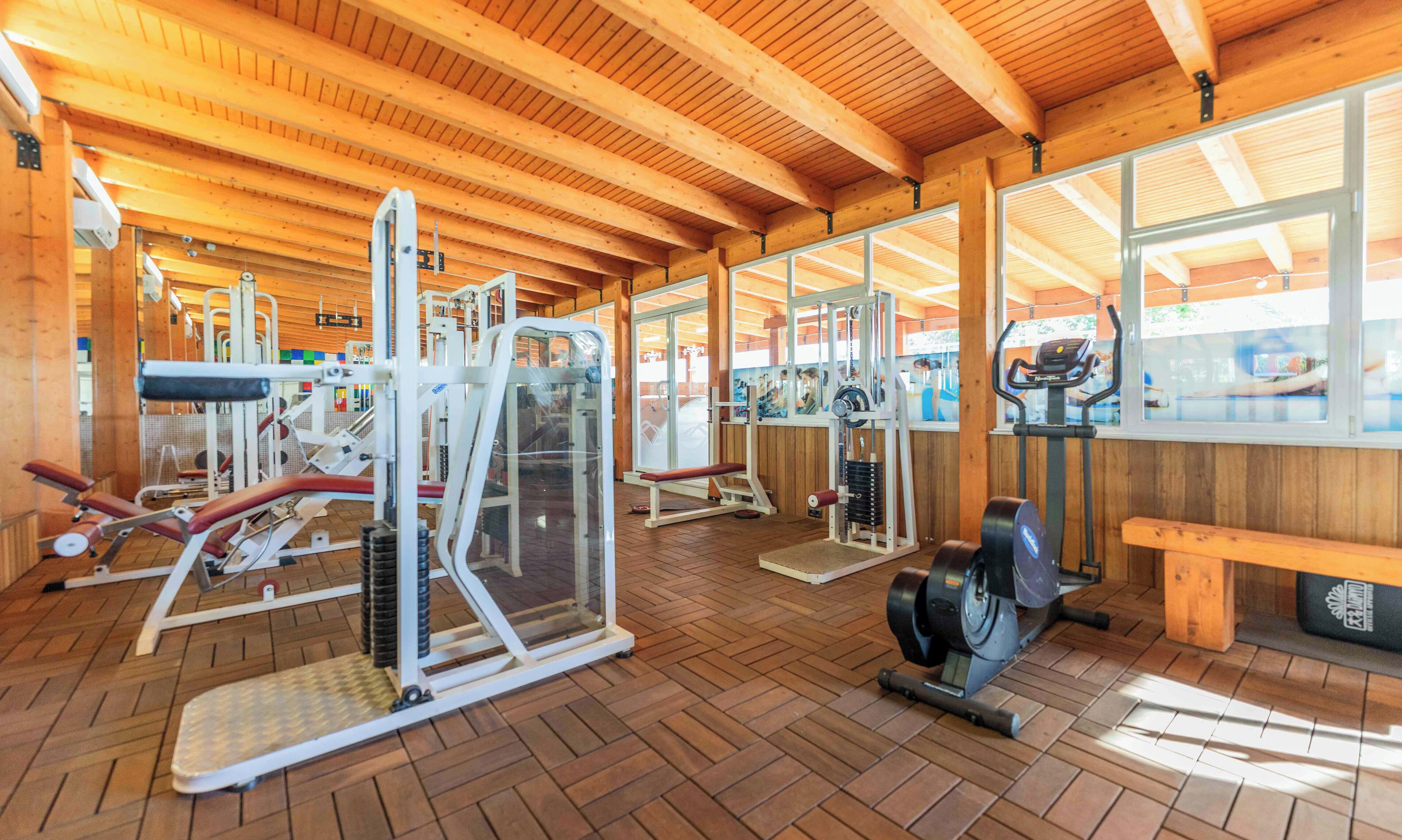 Un gimnasio con diversas máquinas de ejercicio, vigas de madera en el techo y ventanas amplias.