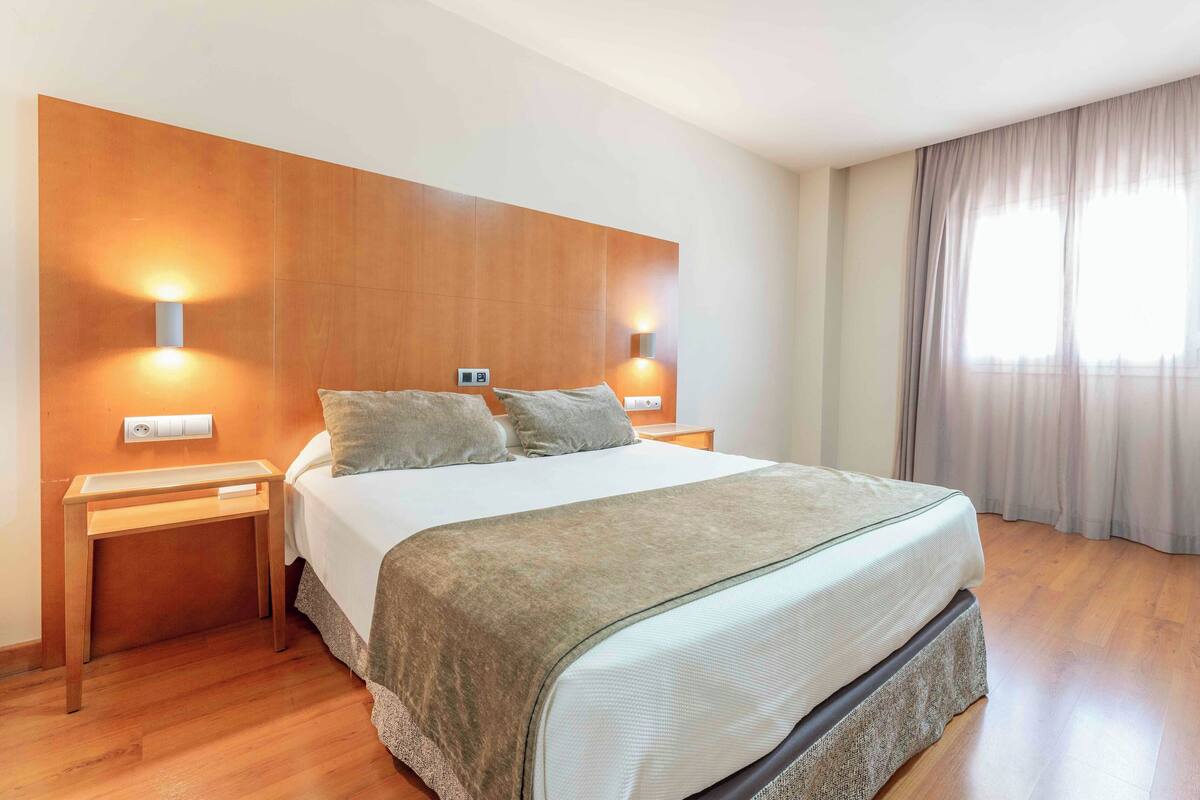 Een moderne hotelkamer met een groot bed, een houten hoofdbord, twee nachtkastjes en een uitzicht op een stadsgezicht door dunne gordijnen.
