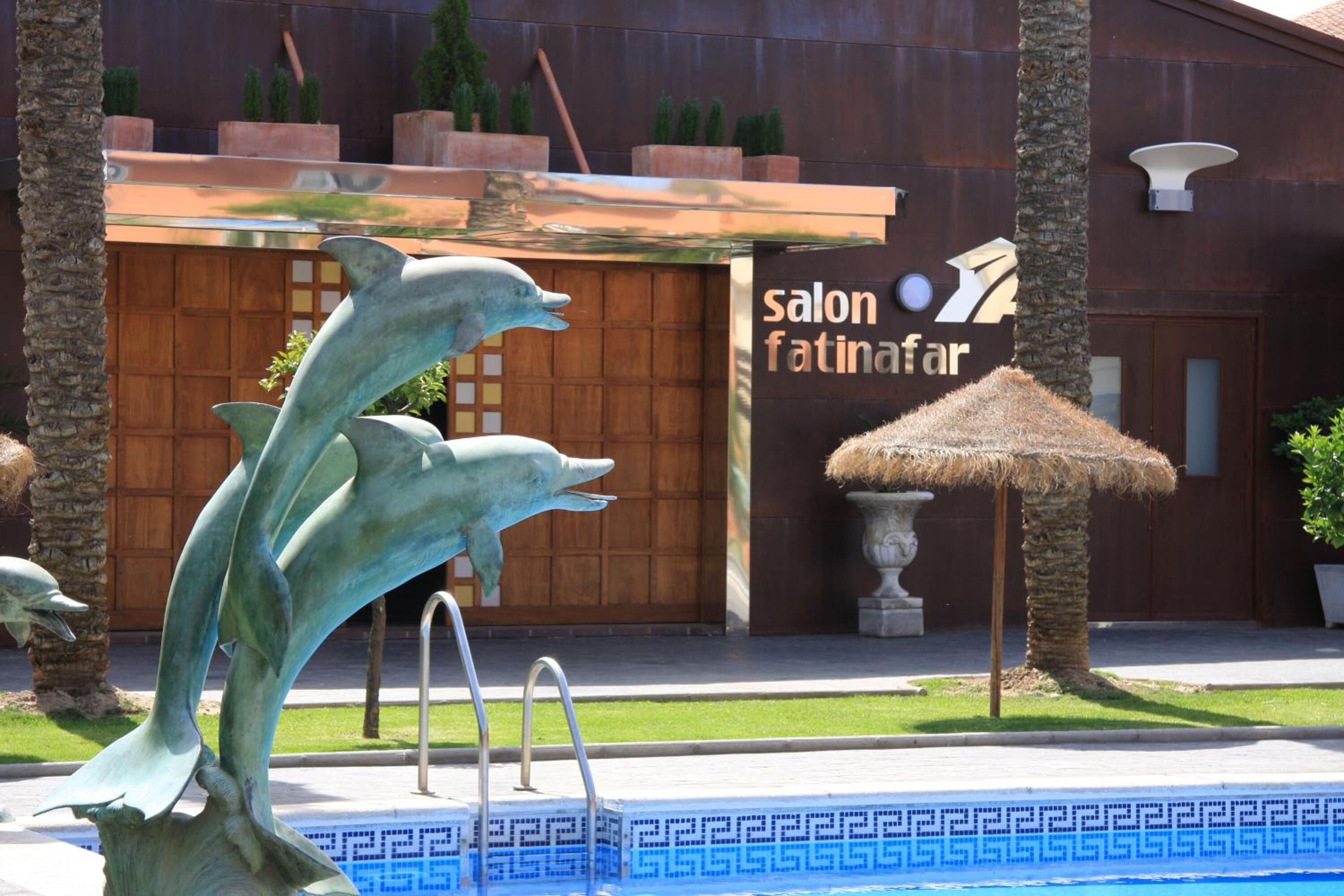Una piscina con una escultura de delfín, un letrero que dice 'Salon Fatinarf' y una sombrilla de paja.