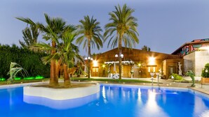 A pool area with palm trees, a building with warm lighting, and a fountain. - Checkin Camino de Granada (Granada)