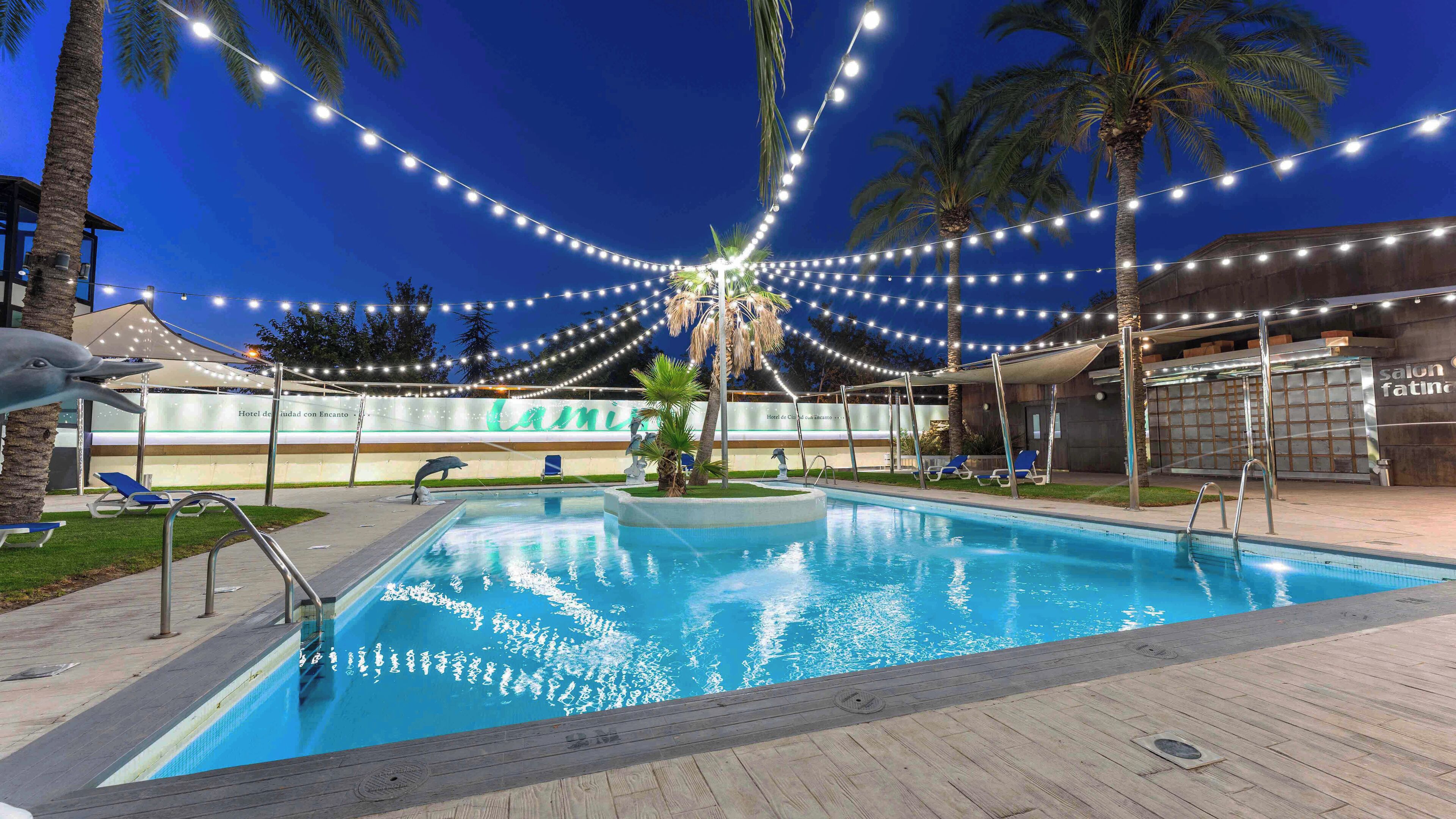 A swimming pool with a wooden deck, lounge chairs, palm trees, and string lights.