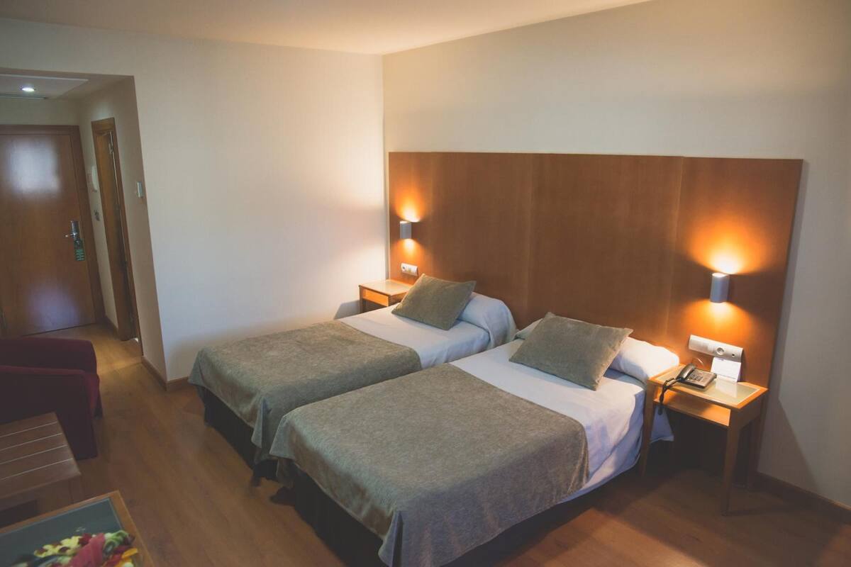 Een hotelkamer met twee bedden, houten hoofdborden, bedlampjes, een bureau en een stoel.