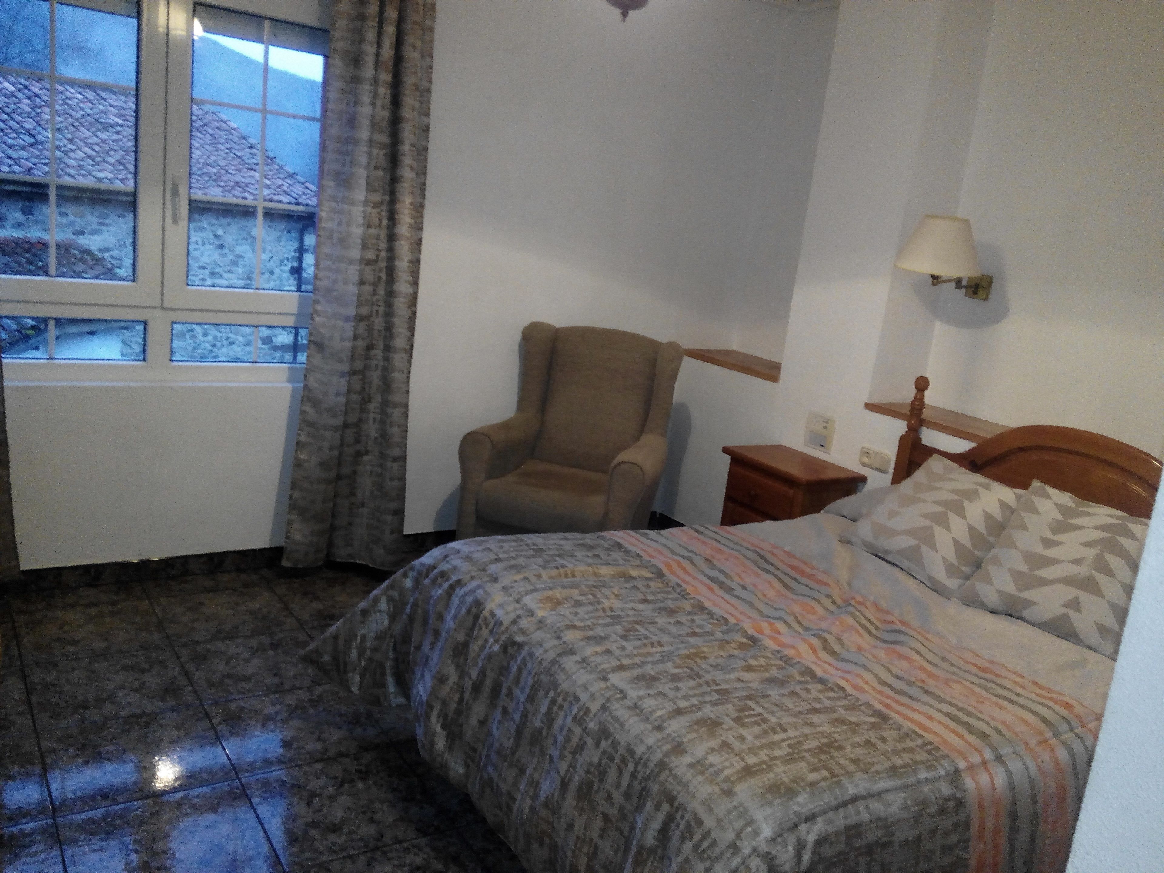 Un dormitorio con cama, una silla, una mesita de noche, una lámpara y una ventana con cortinas.
