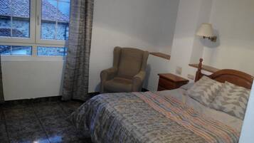 Un dormitorio con cama, una silla, una mesita de noche, una lámpara y una ventana con cortinas.