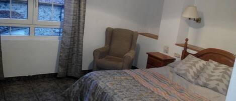 Quarto com cama, cadeira, mesa de cabeceira, luminária e janela com cortinas.