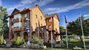 Un edificio de dos pisos con letrero de restaurante, arcos de piedra y un jardín con mesas al aire libre.