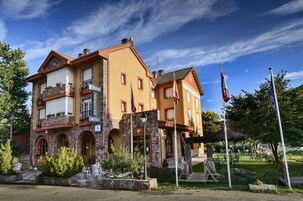 Un edificio de dos pisos con letrero de restaurante, arcos de piedra y un jardín con mesas al aire libre.
