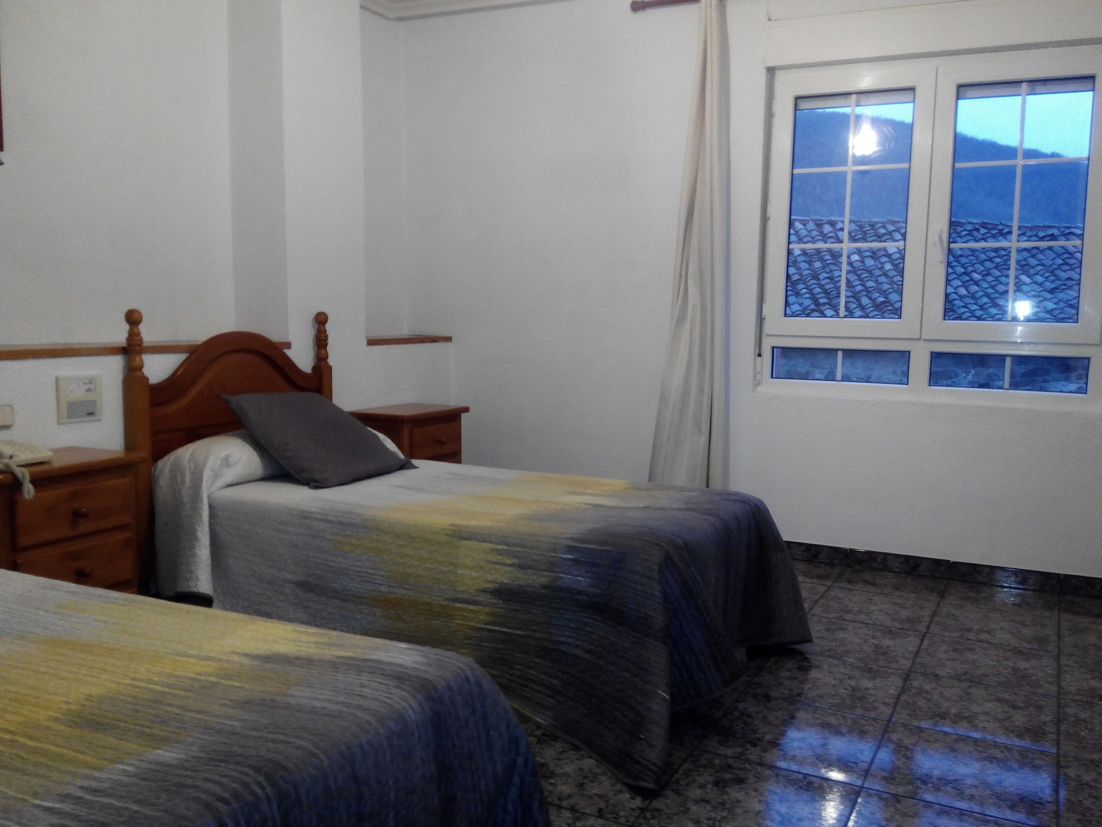 Un dormitorio con dos camas, cabecera de madera, mesita de noche, ventana con vistas a un tejado de tejas y un termostato en la pared.