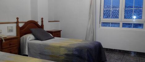 Un dormitorio con dos camas, cabecera de madera, mesita de noche, ventana con vistas a un tejado de tejas y un termostato en la pared.