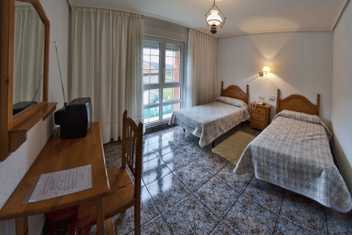 Une chambre avec deux lits, un bureau avec une chaise, un miroir, une fenêtre avec des rideaux et une table de chevet avec une lampe.