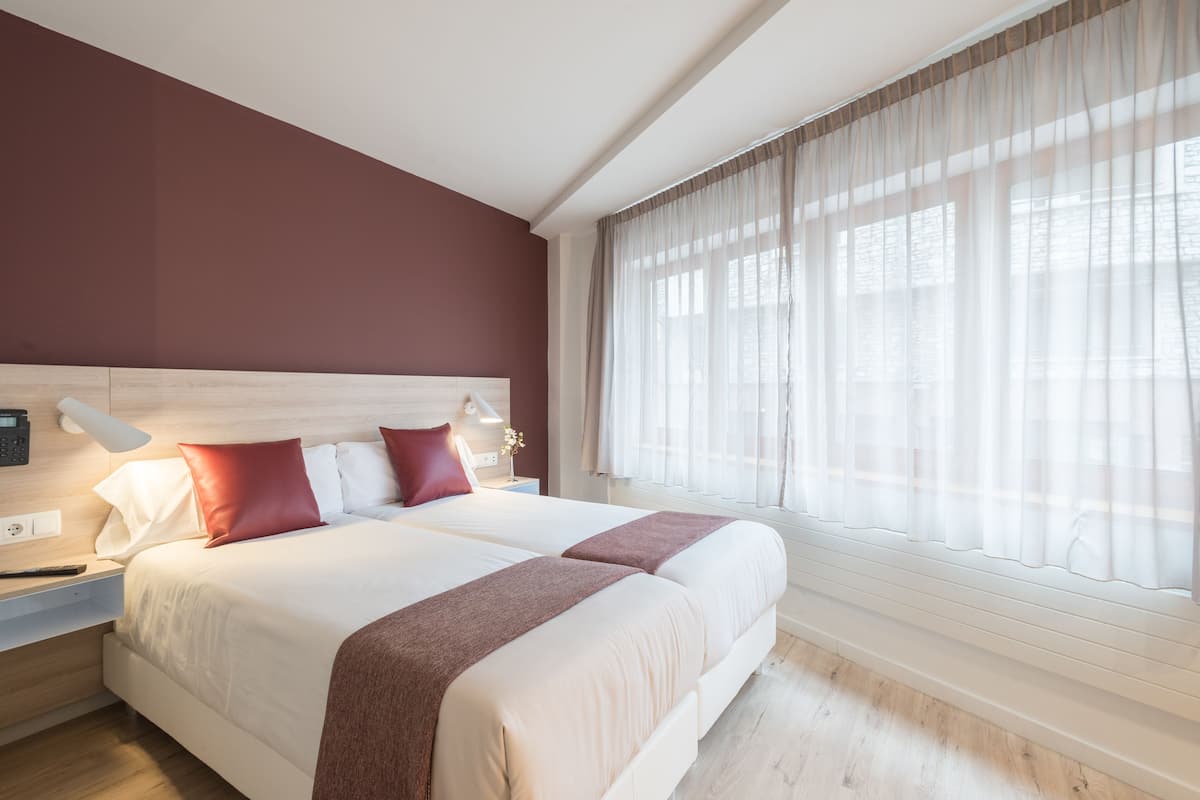 A double bed with white and red bedding, a bedside table with a lamp, and a window with sheer curtains.