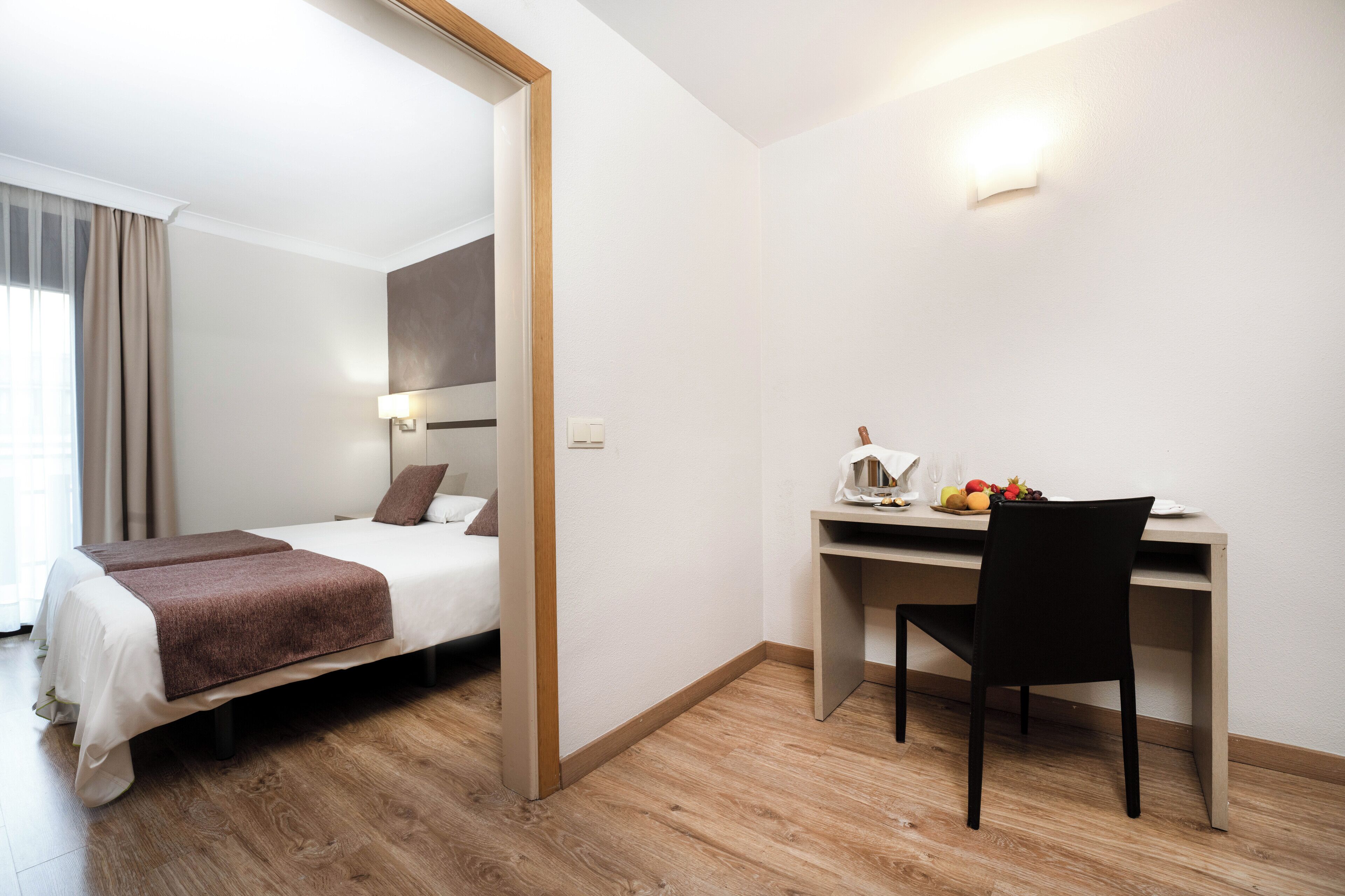 A hotel room with a bed, desk, chair, and a small table with fruits.