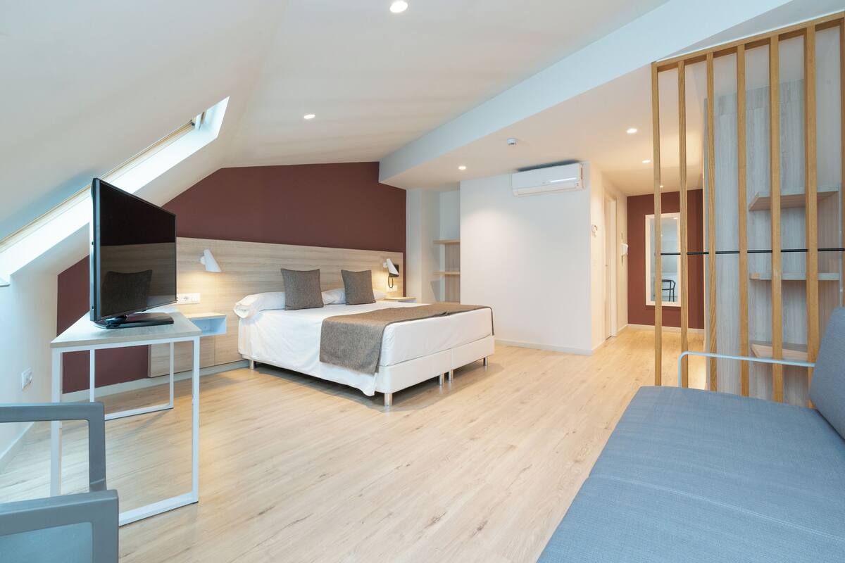 A modern bedroom with a bed, bedside table, and a TV. There is a skylight above the bed and a wooden partition on the right.