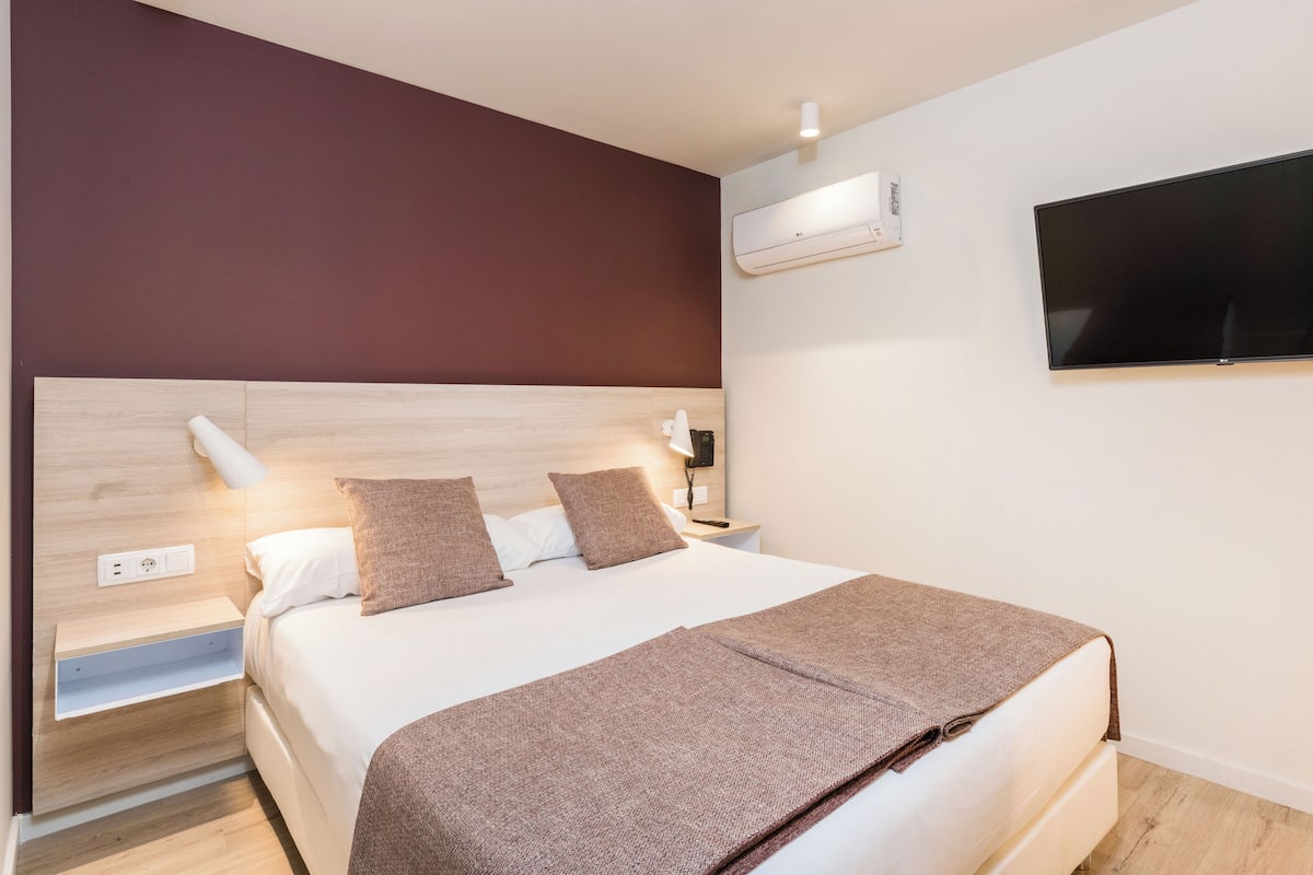 A modern hotel room with a large bed, a flat-screen TV mounted on the wall, a bedside table with a lamp, and an air conditioning unit.