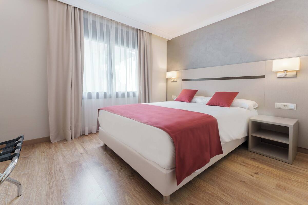 A modern hotel room with a large bed, a bedside table, a bench, and a window with curtains.
