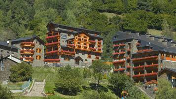 A multi-story hotel with balconies adorned with red flowers, set against a backdrop of lush green hills.