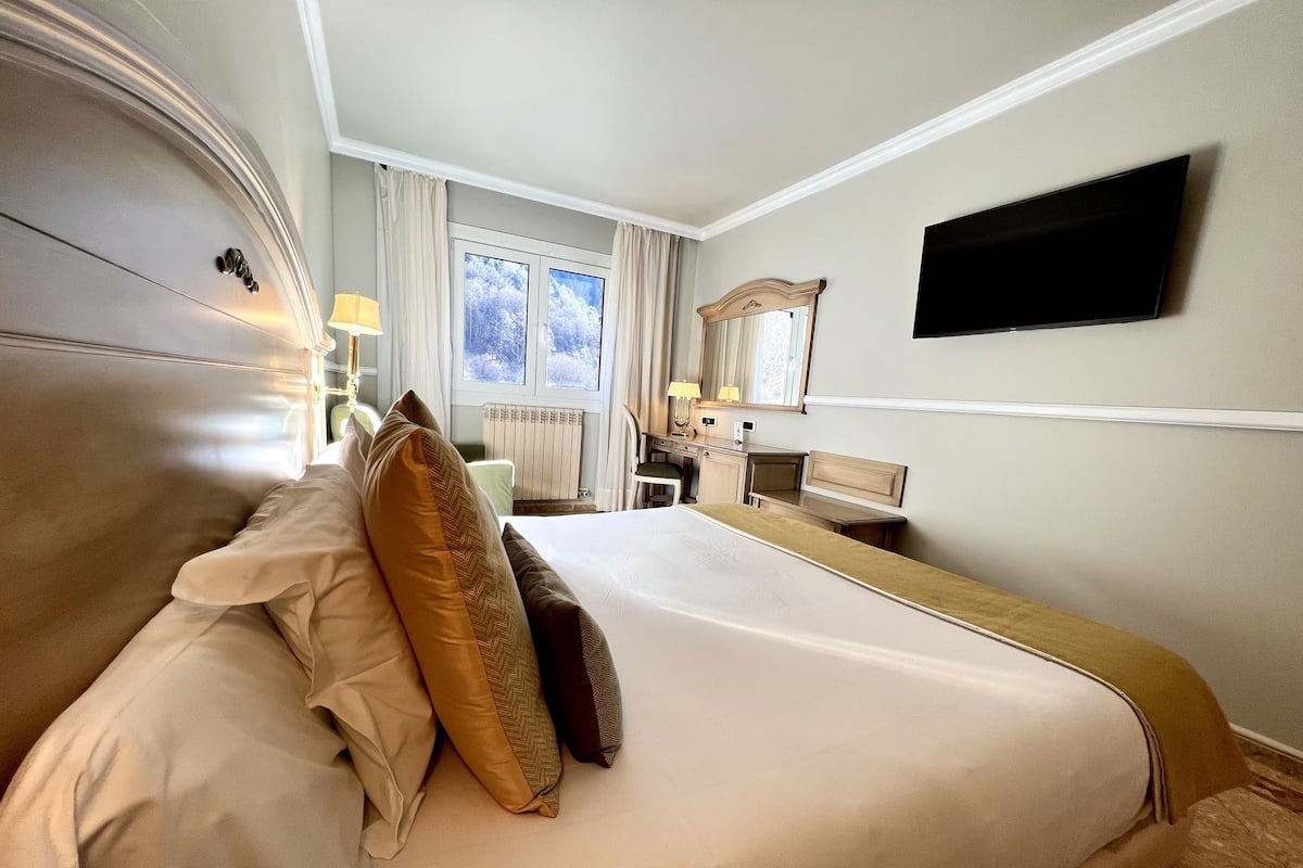 A hotel room with a large bed, a desk, a chair, a TV mounted on the wall, and a window with curtains.