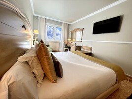 Una habitación de hotel con una cama grande, un escritorio, una silla, un televisor montado en la pared y una ventana con cortinas.
