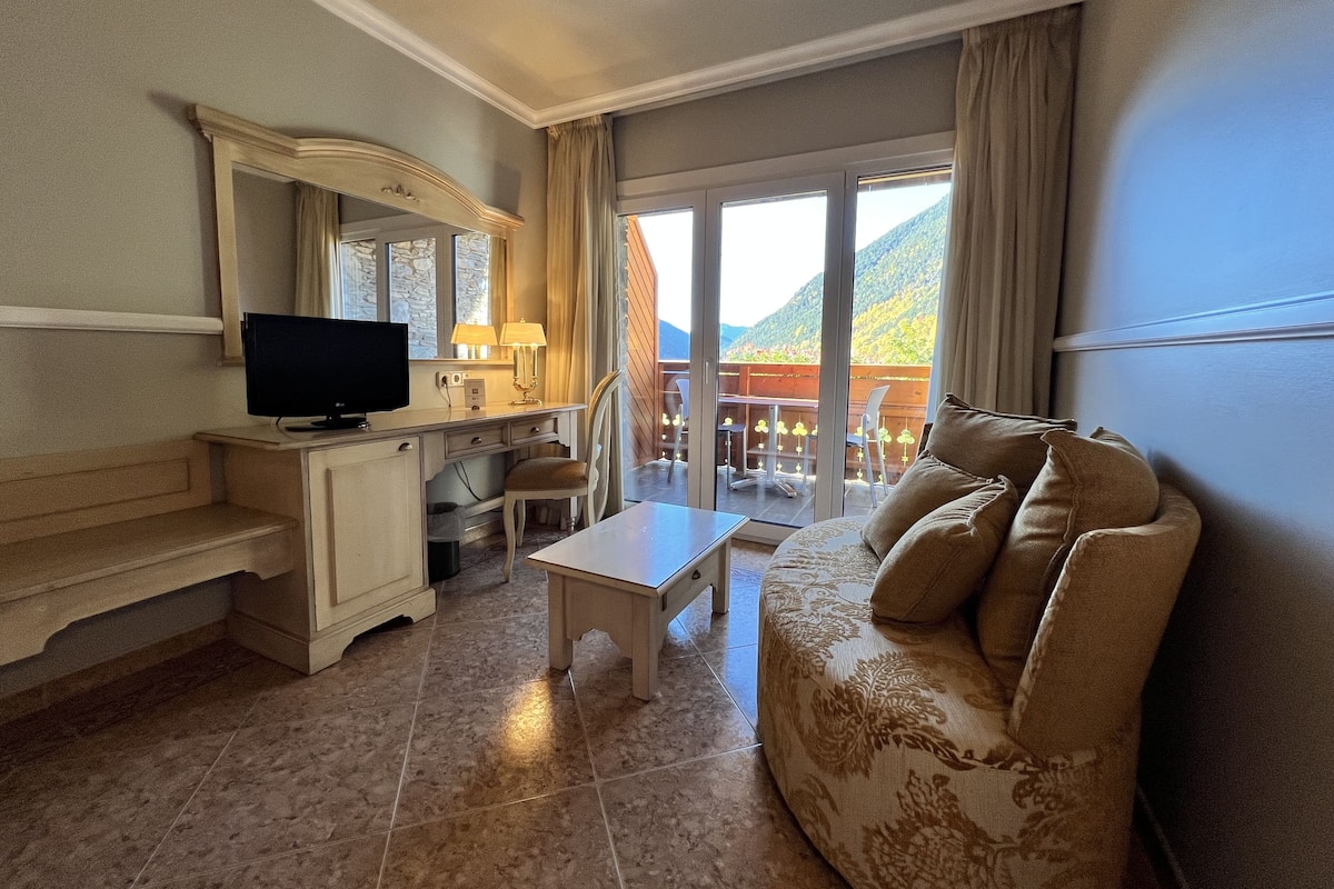 A hotel room with a sofa, a TV, a desk, and a balcony with a mountain view.