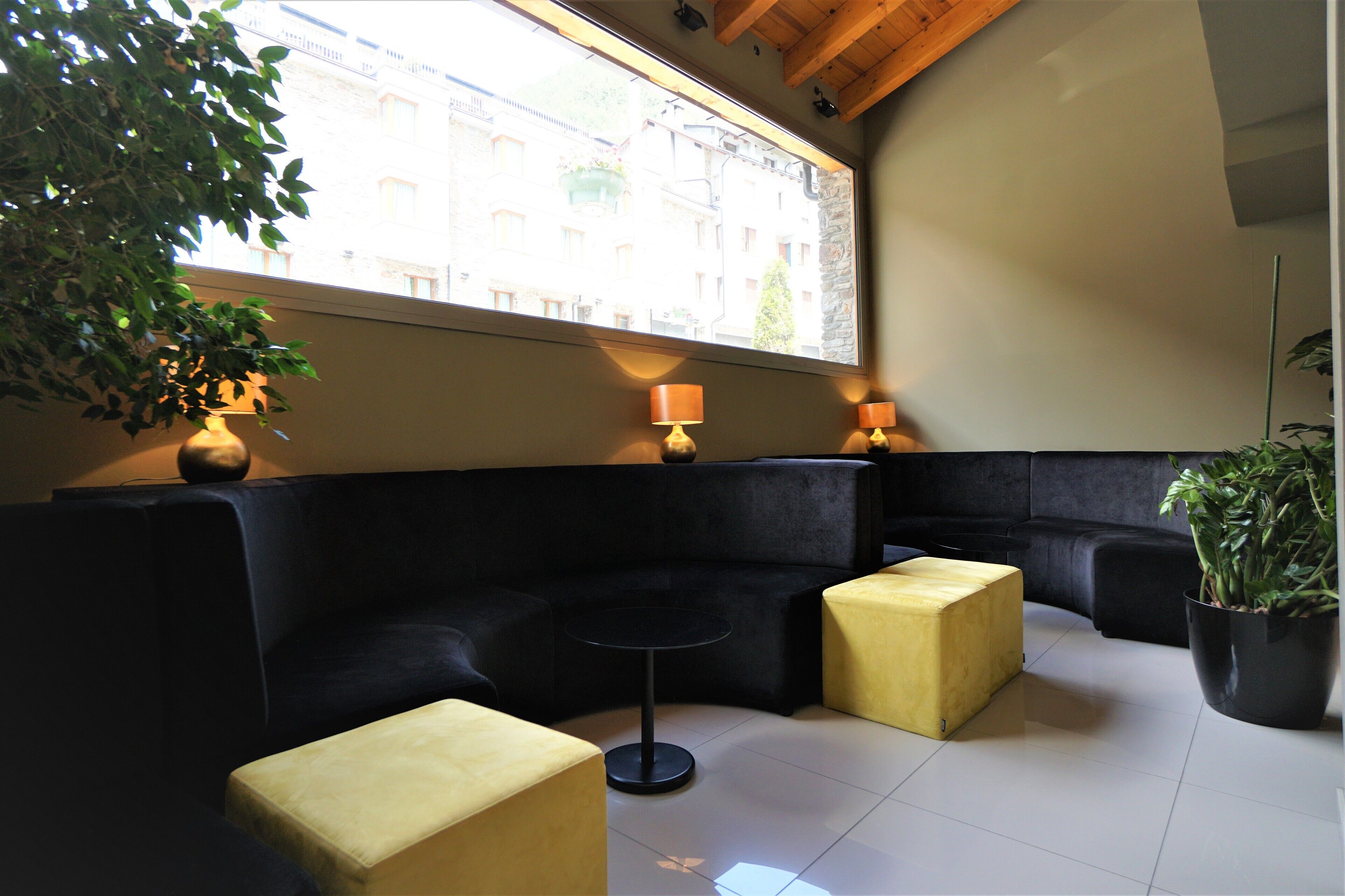 A modern lounge area with a black sofa, a small round table, and two yellow ottomans. A large window provides a view of buildings outside.