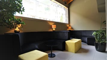 A modern lounge area with a black sofa, a small round table, and two yellow ottomans. A large window provides a view of buildings outside.