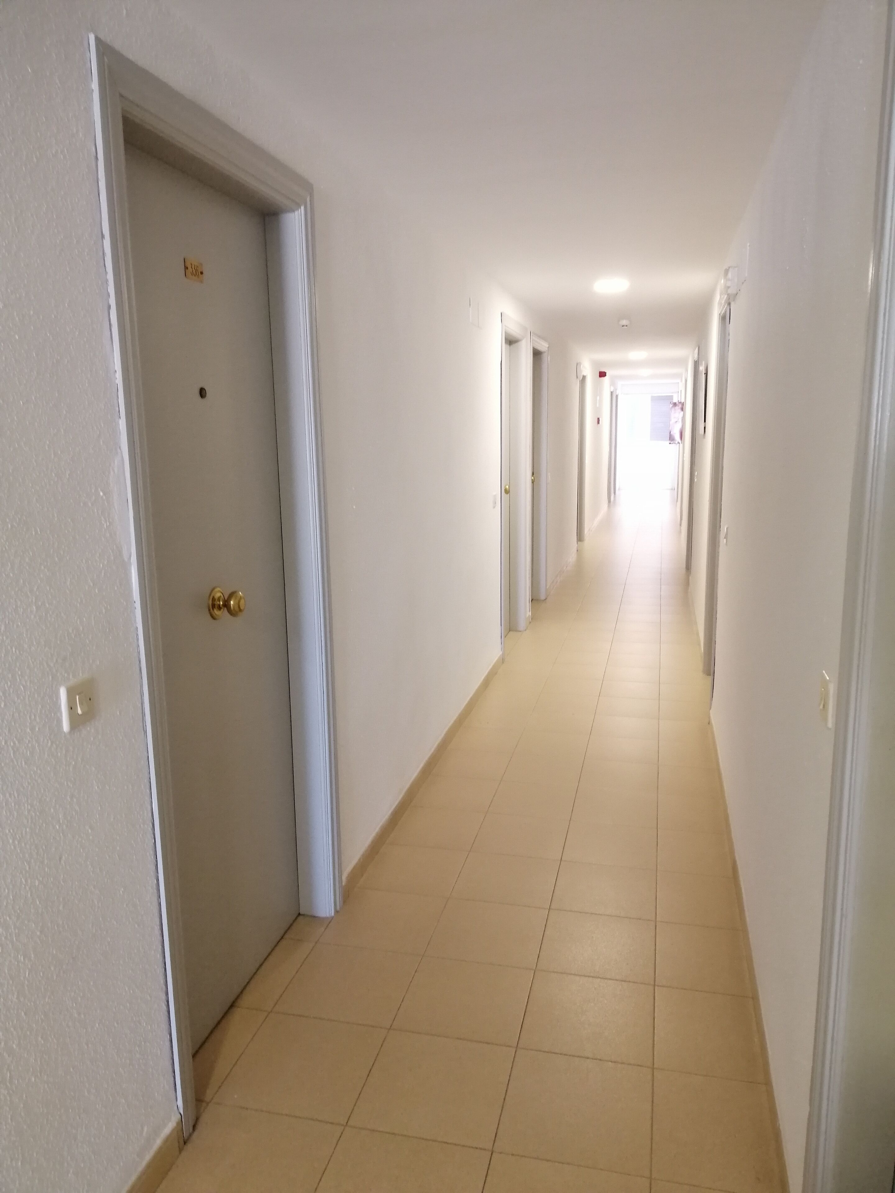 A long, narrow hallway with white walls and tiled floors, leading to multiple doors.