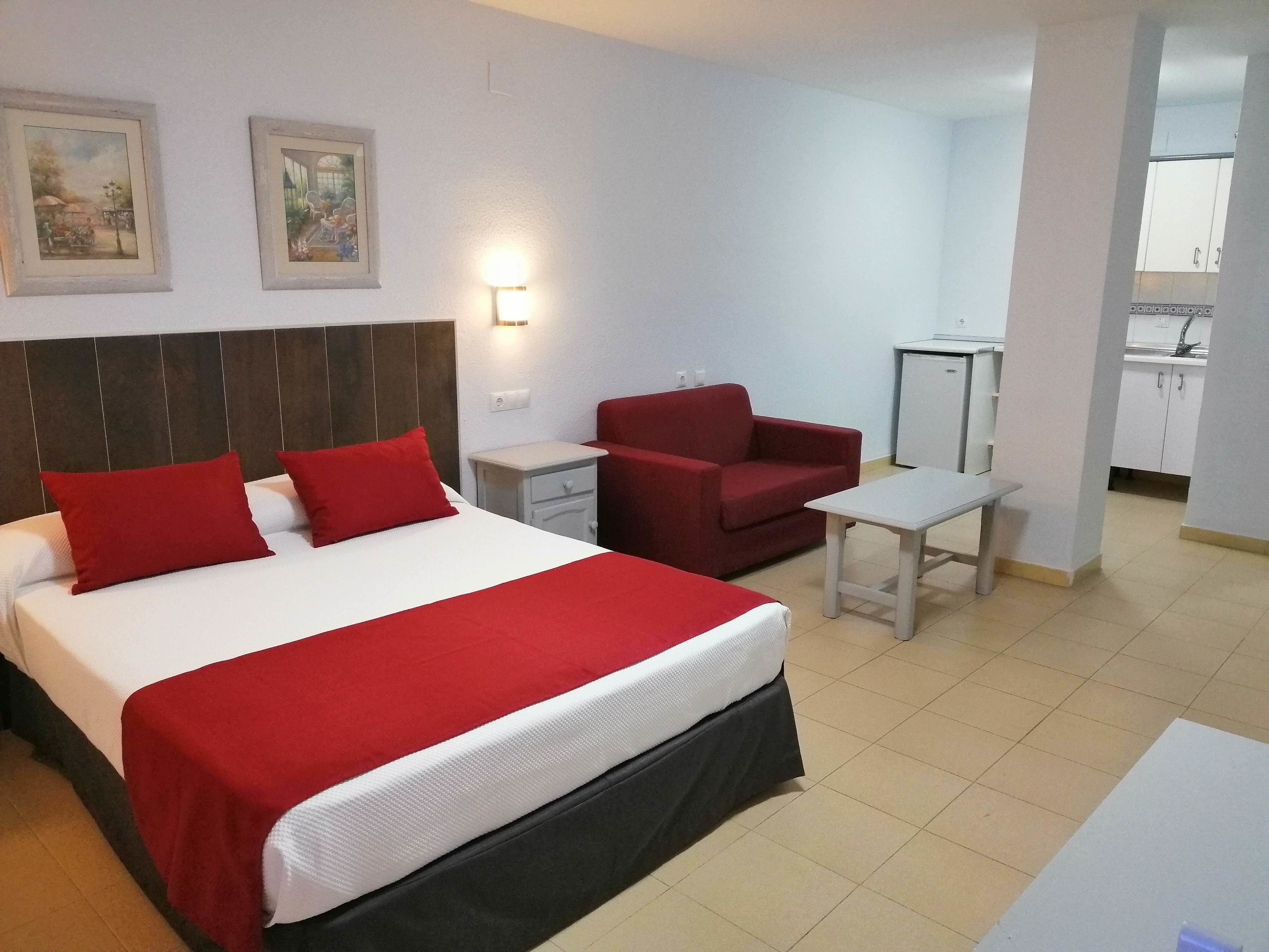 Una habitación de hotel con cama, una silla roja, una mesita y una kitchenette.