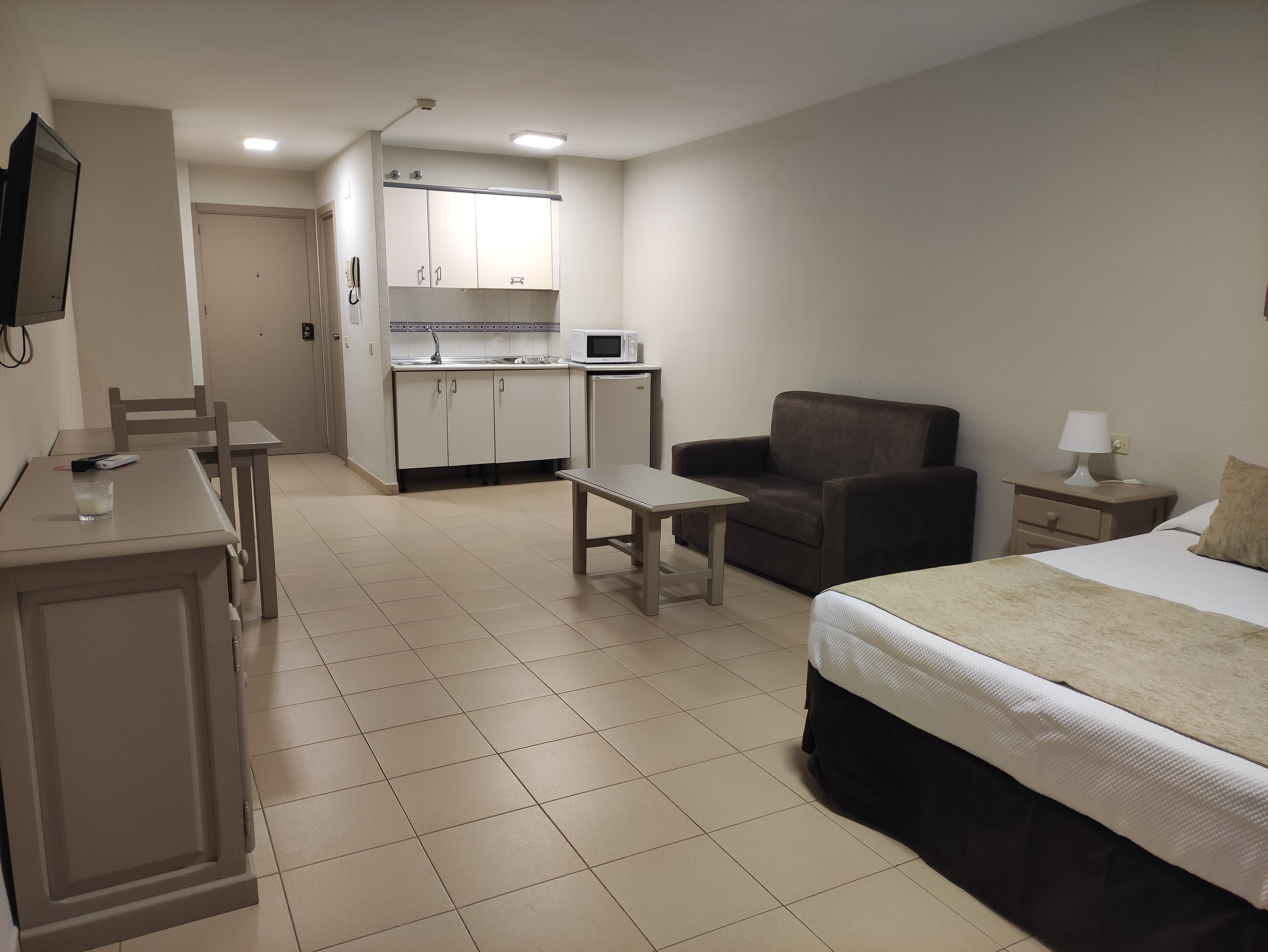 A compact hotel room with a bed, a small desk, a chair, a sofa, and a kitchenette.
