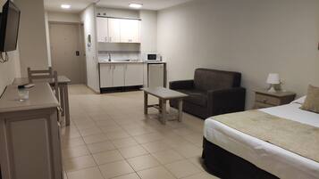A compact hotel room with a bed, a small desk, a chair, a sofa, and a kitchenette.