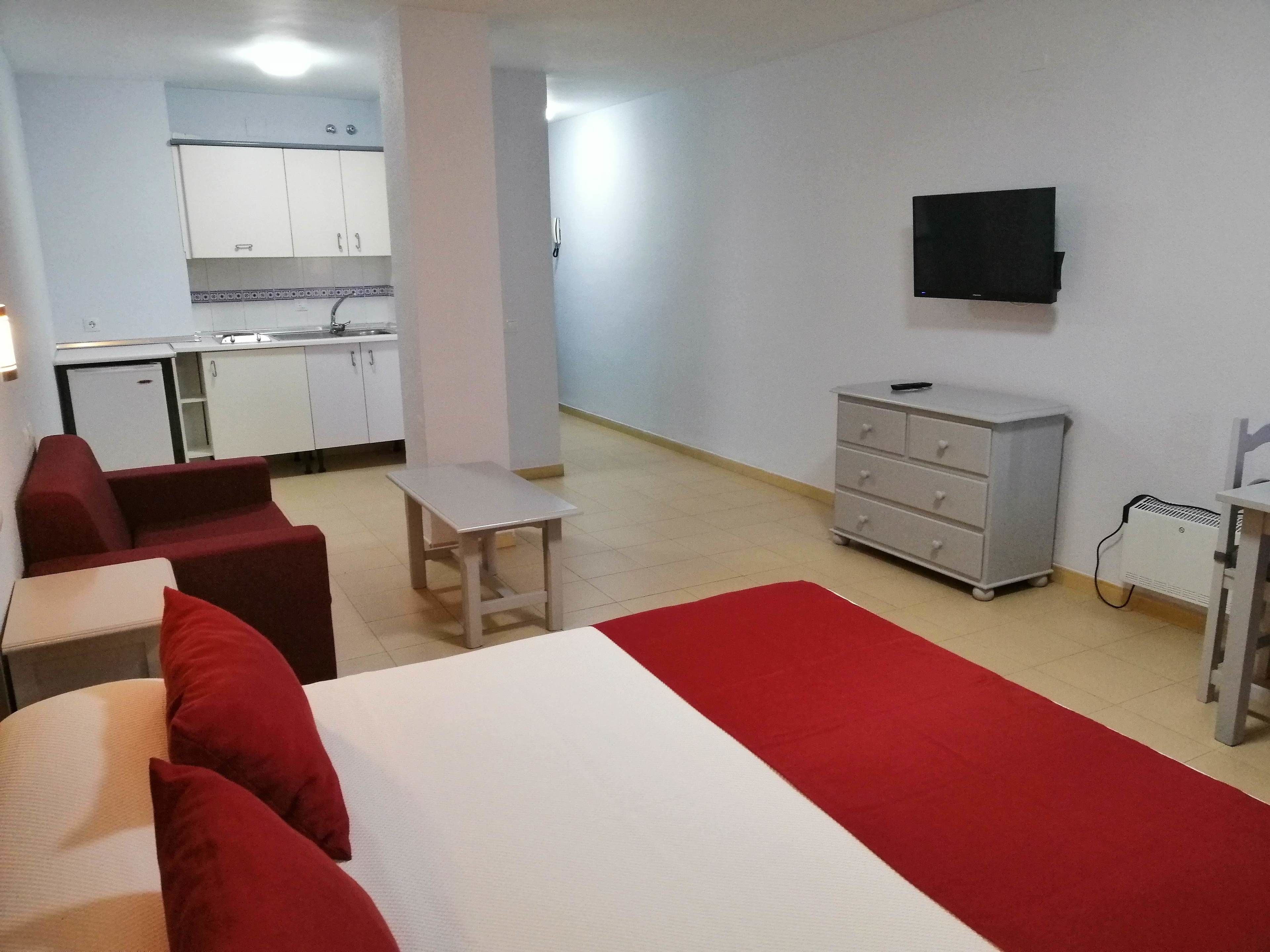 A compact hotel room with a bed, a small table, a sofa, a kitchenette, and a TV mounted on the wall.