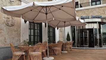 An outdoor seating area with wicker chairs and large umbrellas, a stone wall, and a building entrance with a sign reading "CONDE ANSUREZ".