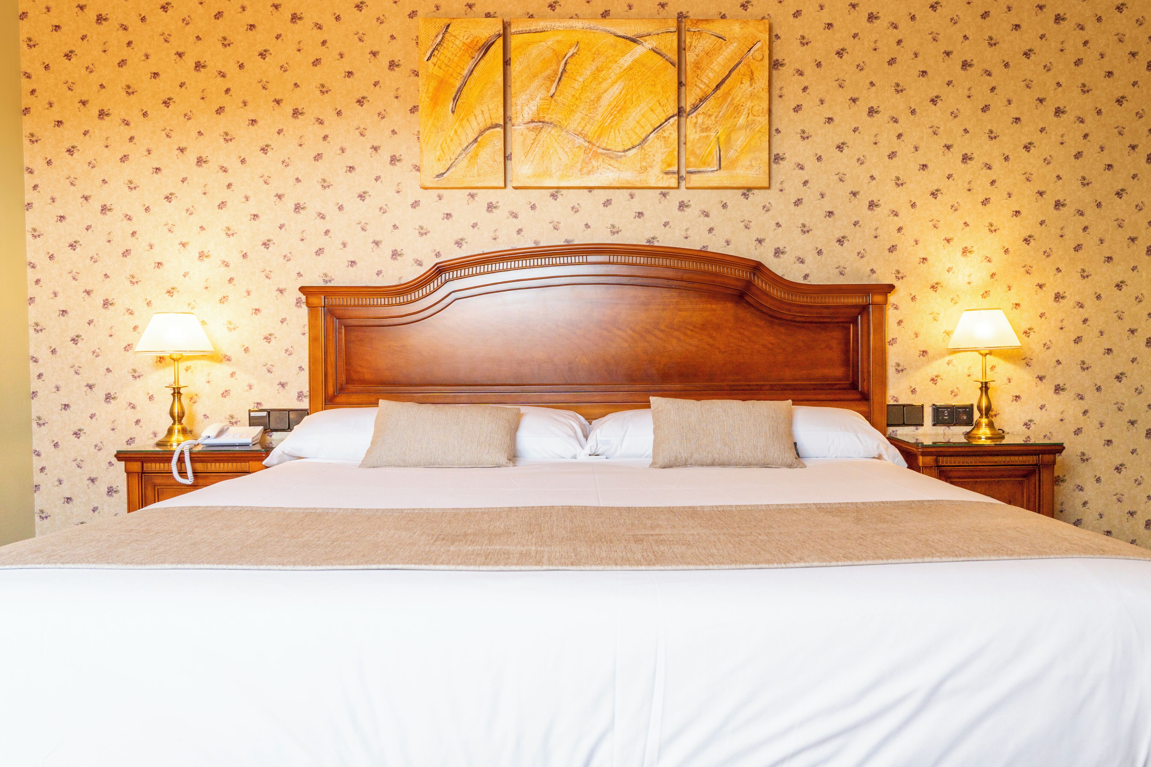 Een net opgemaakt bed met een houten hoofdbord, twee bedlampen en een behang met een bloemendesign.