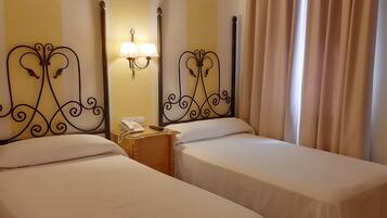 A hotel room with two single beds, a bedside table with a phone and a lamp, and a curtain on the window.