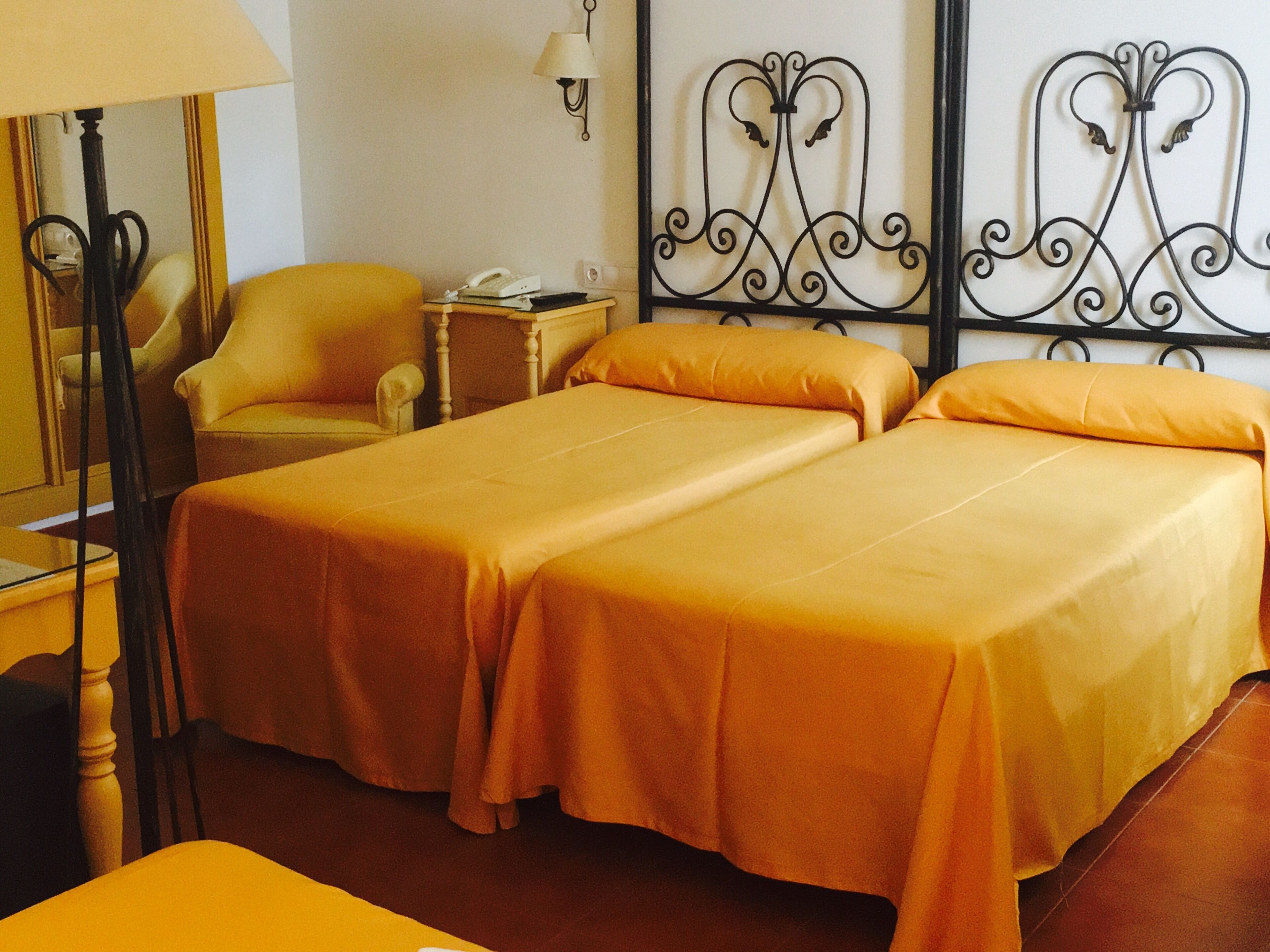 Habitación de hotel con dos camas individuales, una silla, una mesita de noche con lámpara y un adorno metálico en la pared.