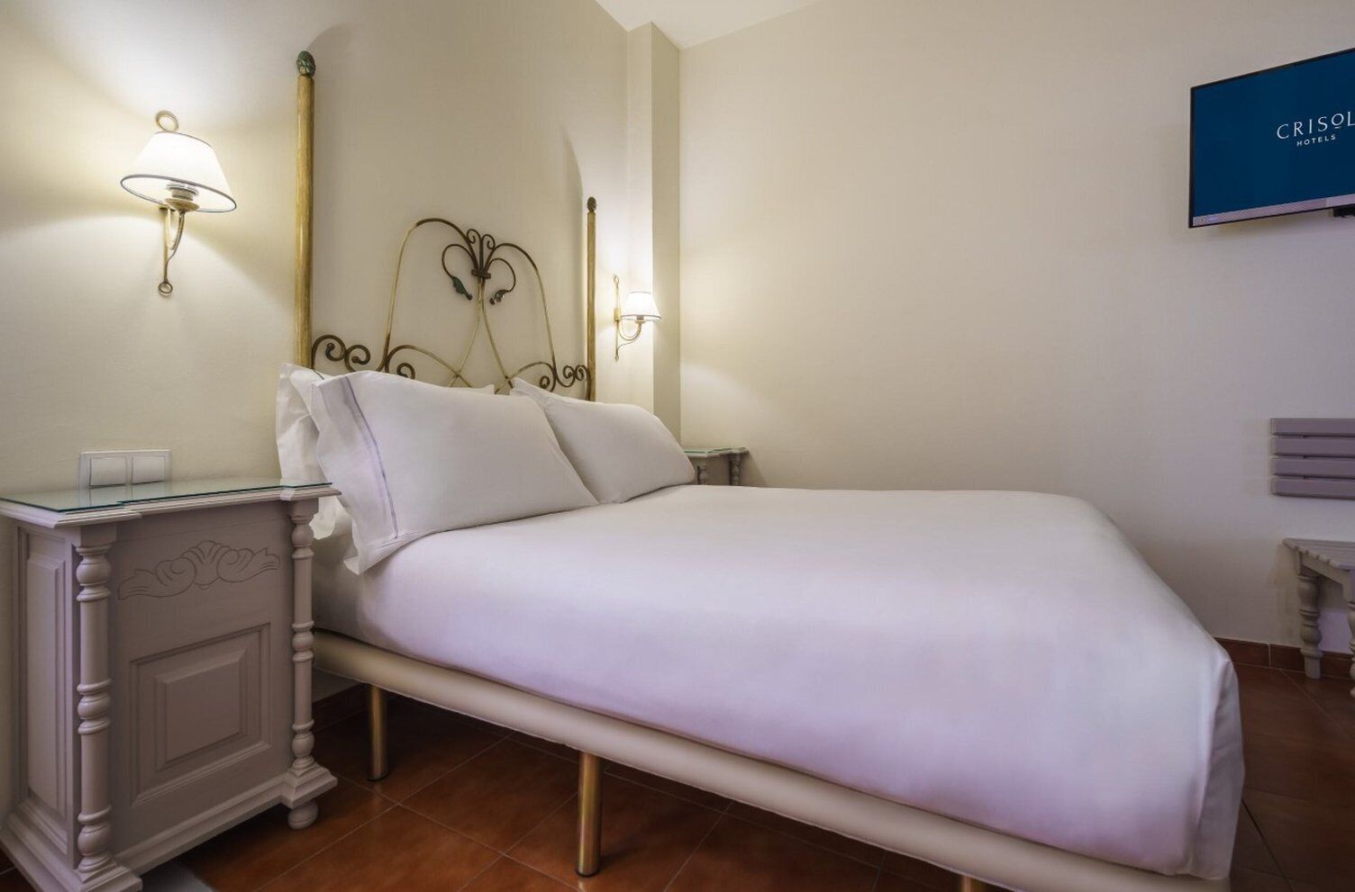 Cama bem arrumada com edredom branco e almofadas, mesa de cabeceira com abajur e televisão fixada na parede.