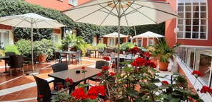 Een terras buiten met meerdere tafels en stoelen, omgeven door potplanten en rode bloemen.