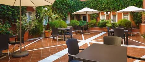 An outdoor patio area with tiled flooring, tables, chairs, and umbrellas.